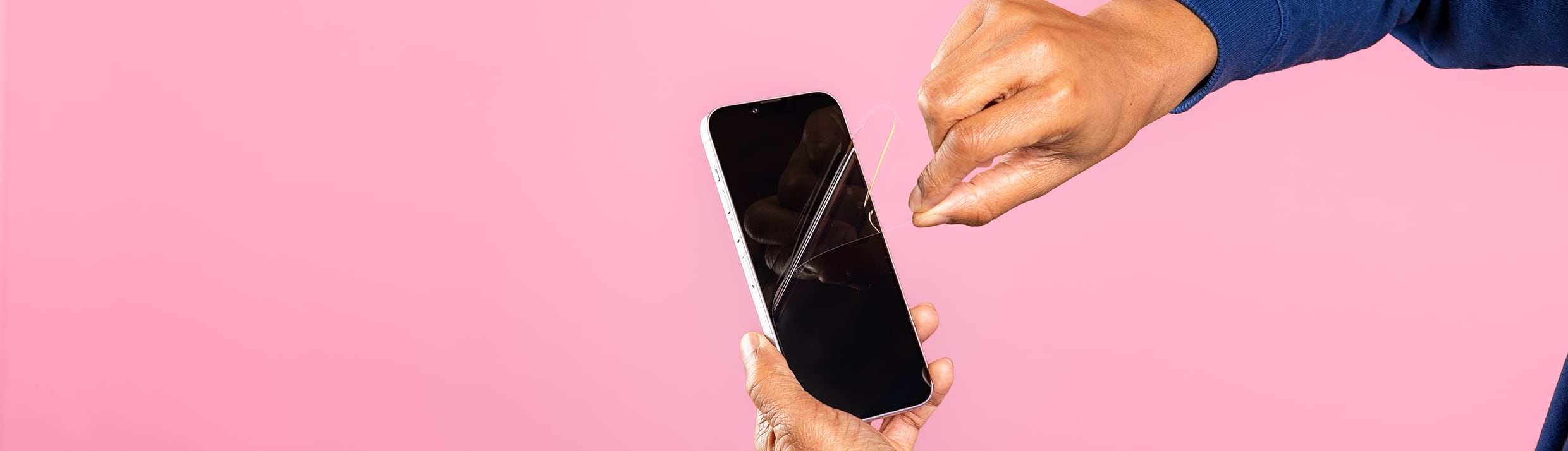 Deux mains retirent un film protecteur d'un iPhone, l'arrière-plan est rose.