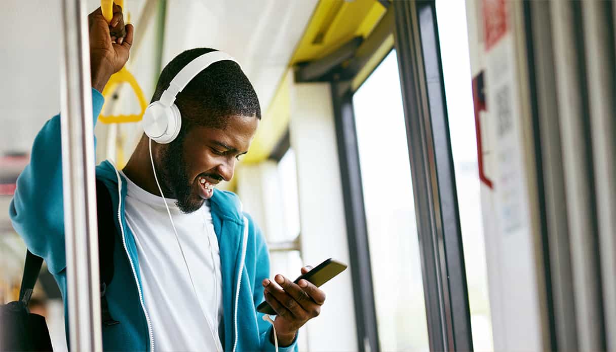 Un homme est debout dans le bus et regarde son t&eacute;l&eacute;phone en riant, il porte un casque avec fil.