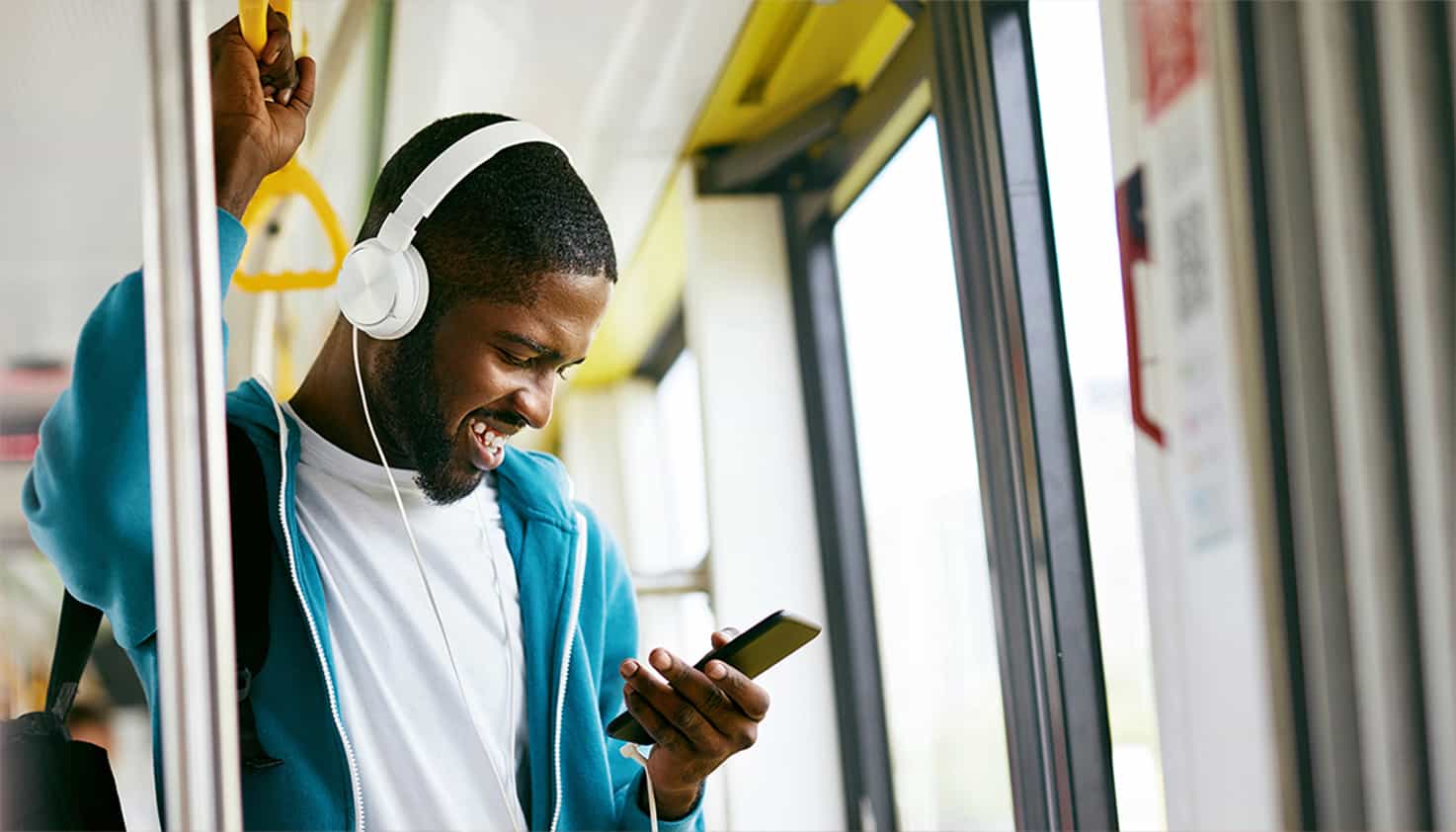Un homme est debout dans le bus et regarde son t&eacute;l&eacute;phone en riant, il porte un casque avec fil.