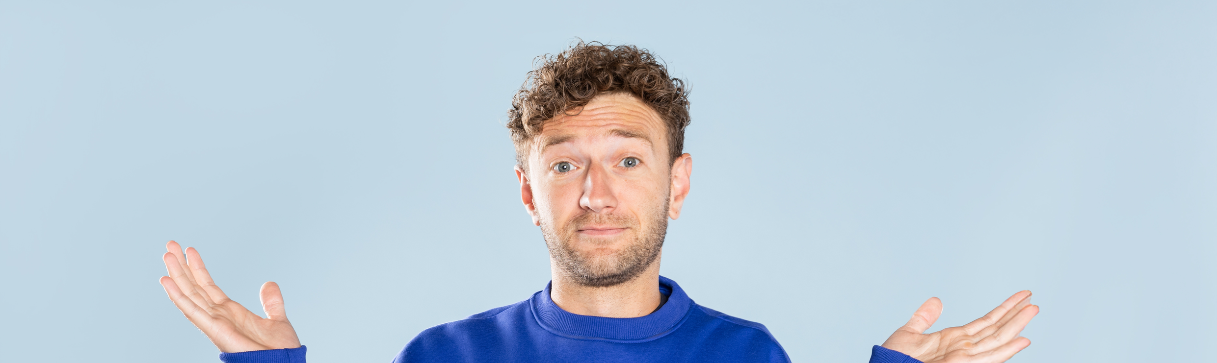 Man in blauwe trui die vragend zijn handen omhoog doet.