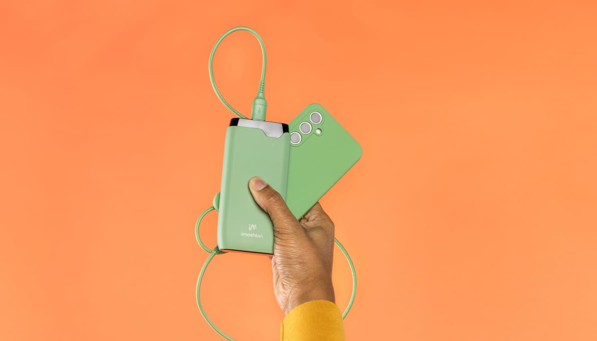 Une main dont on aperçoit un bout de manche jaune sort du bas de l'image et tient un téléphone avec une coque verte, un câble de recharge vert et une batterie externe imoshion verte. L'arrière-plan est orange.