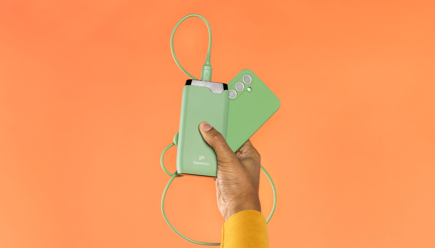 Une main dont on aperçoit un bout de manche jaune sort du bas de l'image et tient un téléphone avec une coque verte, un câble de recharge vert et une batterie externe imoshion verte. L'arrière-plan est orange.