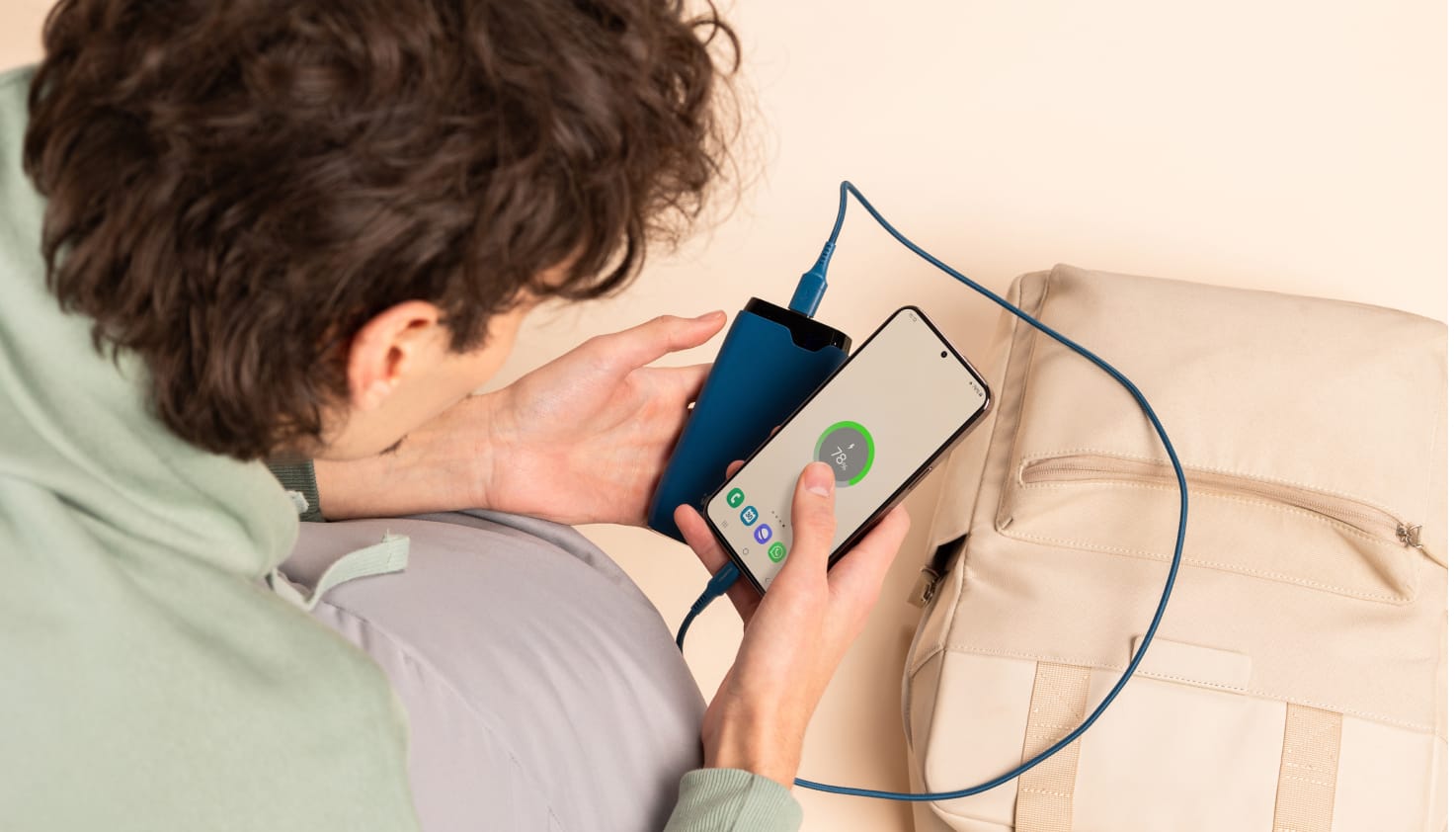 Vous voyez une personne aux cheveux bruns regardant un smartphone qui se recharge via un câble bleu branché sur une batterie externe bleue. Sur le fond beige se trouve un sac à dos couleur sable.