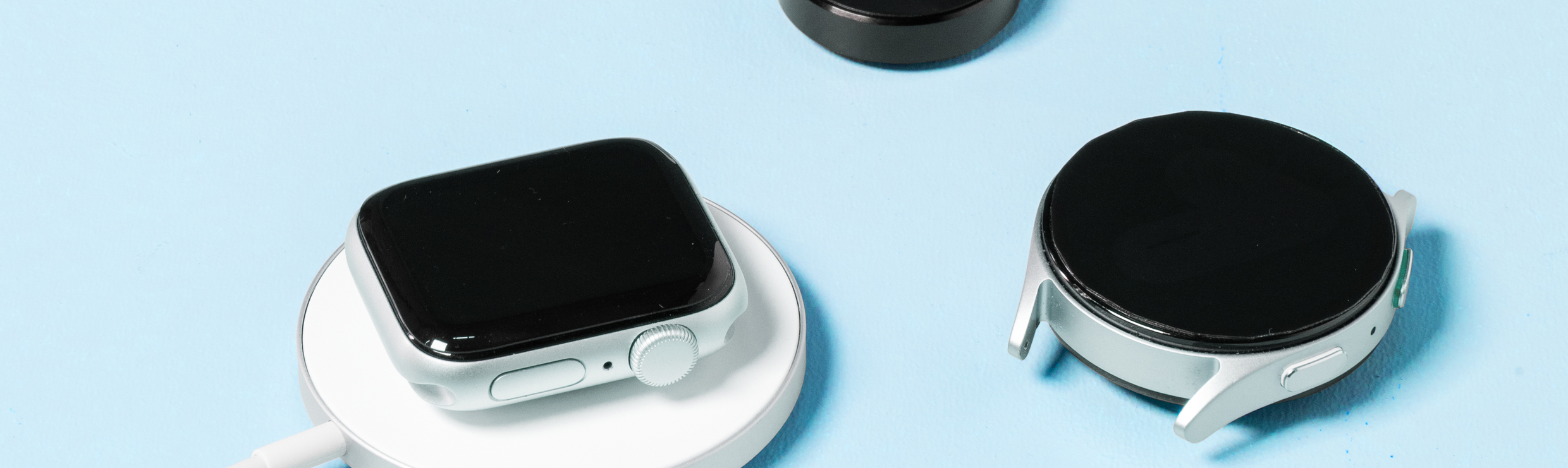 Une Apple Watch sur un chargeur sans fil, à côté se trouvent une Samsung Watch et un chargeur de smartwatch séparé.