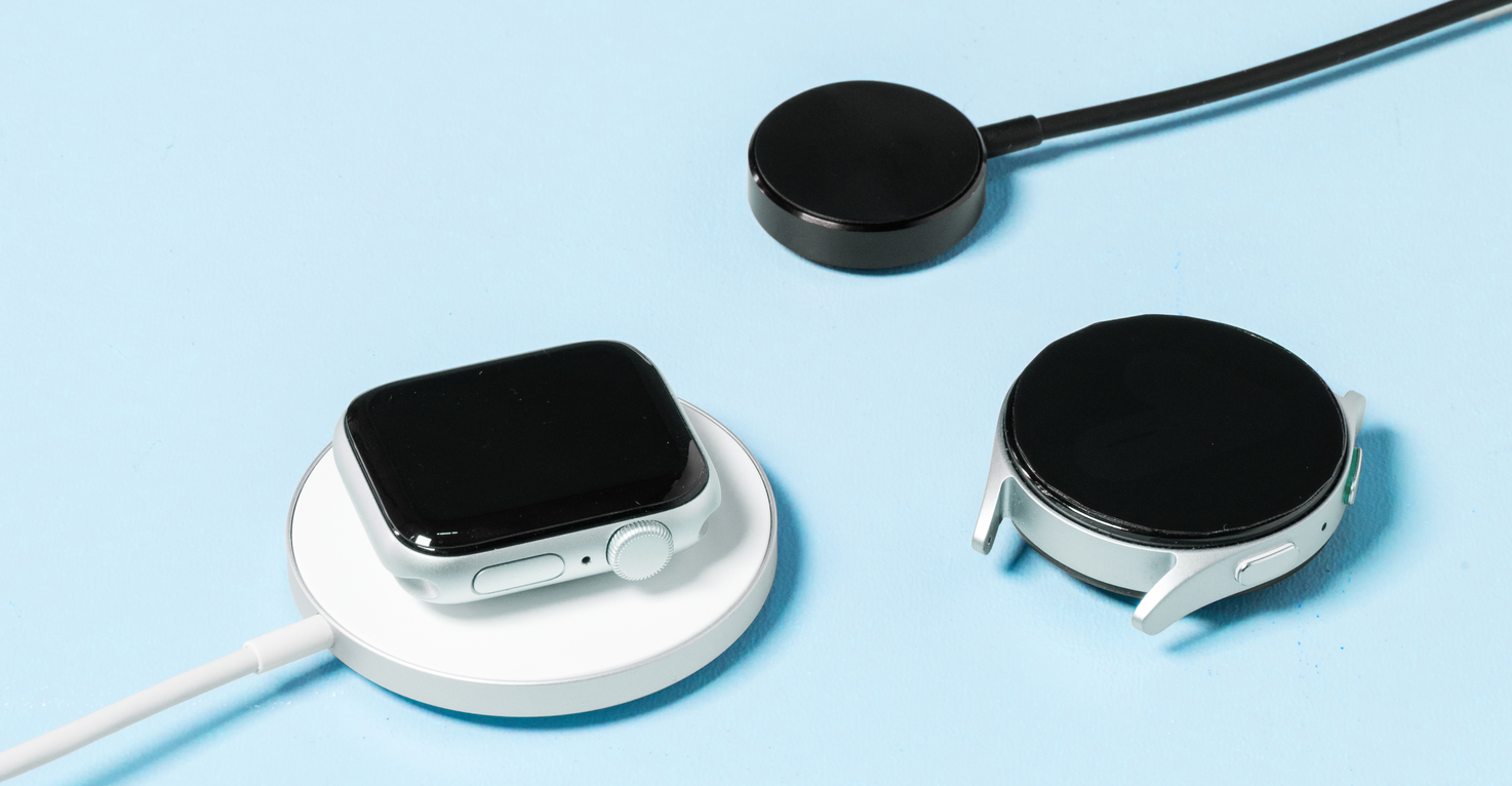 Une Apple Watch sur un chargeur sans fil, à côté se trouvent une Samsung Watch et un chargeur de smartwatch séparé.